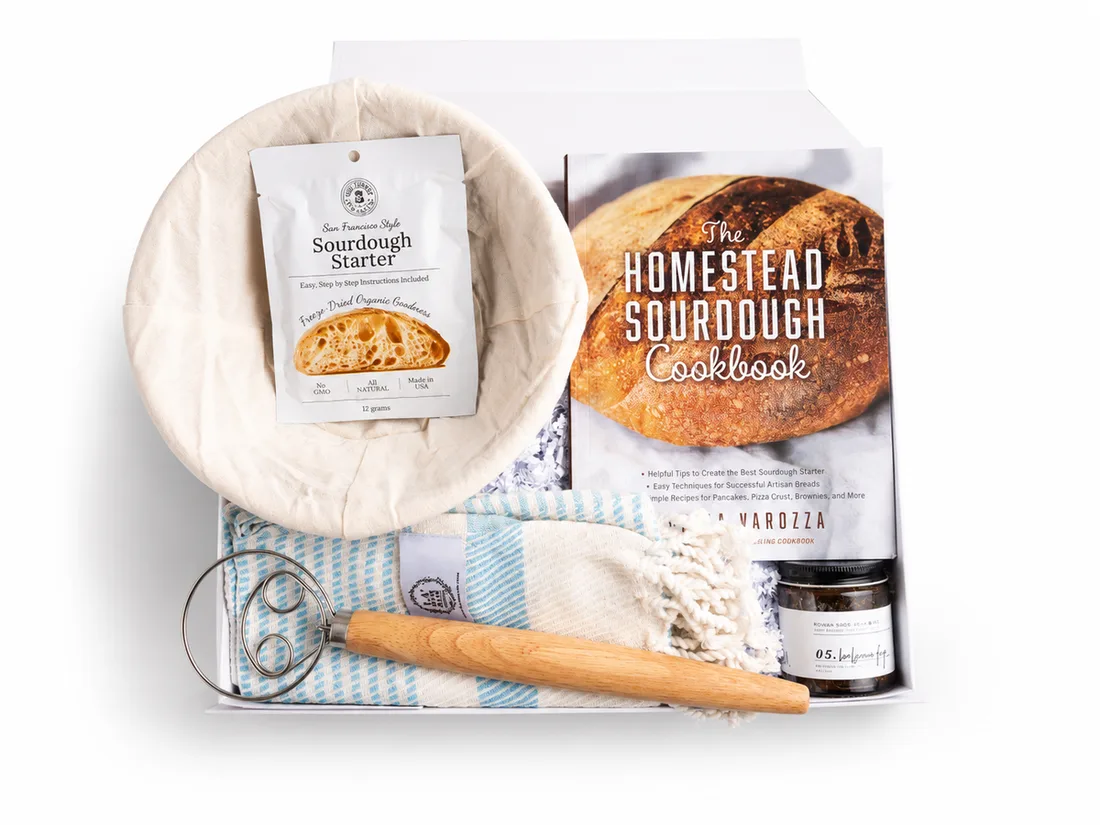 White gift box holding a sourdough starter, proofing basket, cookbook, dough whisk and a striped linen towel