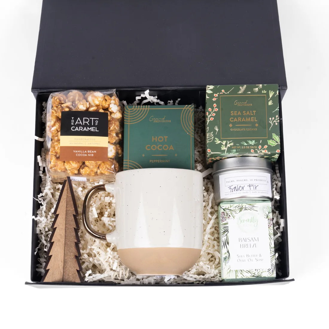 Black gift box holding a speckled mug, hot cocoa, sea-salt caramel cookies, caramel popcorn, a soy candle and a wooden tree ornament