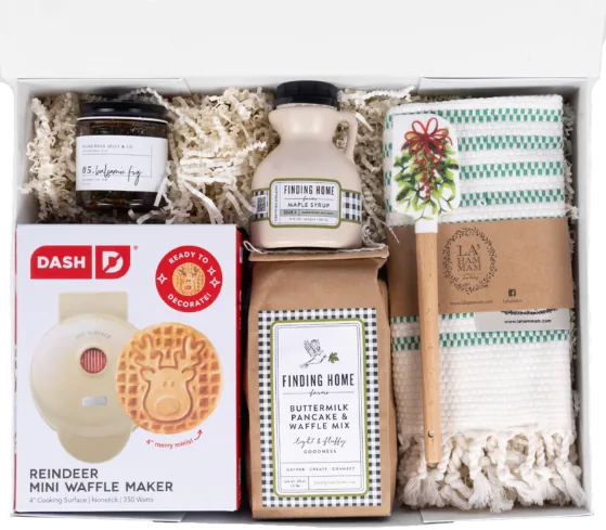 White box holding a reindeer mini waffle maker, buttermilk pancake and waffle mix, maple syrup, fig jam and a striped kitchen towel